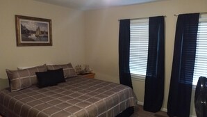 A bedroom with a bed, a nightstand, a lamp, a window with curtains, and a painting on the wall.