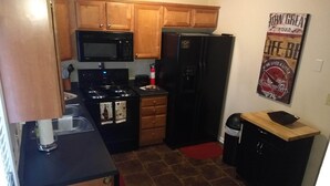 A compact kitchen with wooden cabinets, a black microwave, a refrigerator, and a small trash bin.