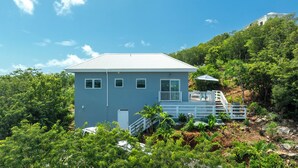 Une maison à deux étages dotée d’un balcon et d’une petite terrasse, entourée d’une végétation luxuriante et d’arbres.