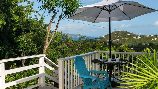 Une terrasse dotée d’une balustrade blanche, d’une chaise bleue, d’une table avec un parasol noir et offrant une vue sur un paysage vallonné.