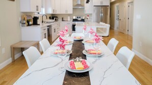 Una cocina moderna con armarios blancos, una isla central y un comedor listo para una comida, con sillas blancas y una mesa de mármol.