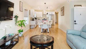 Una cocina moderna con armarios blancos, un comedor con mesa y sillas blancas, un sofá azul y un televisor montado en la pared.