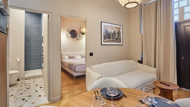 Ein modernes Hotelzimmer mit einem Holzesstisch für zwei Personen, einer Couch, einem Bett und einem Badezimmer mit einer Duschkabine mit blauen Fliesen.