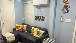 Ein Wohnzimmer mit einem dunkelgrauen Ecksofa, bunten Kissen, einem kleinen gestreiften Hocker, einem flauschigen Teppich, einem an der Wand befestigten Kleiderhaken und einem dekorativen Gecko.