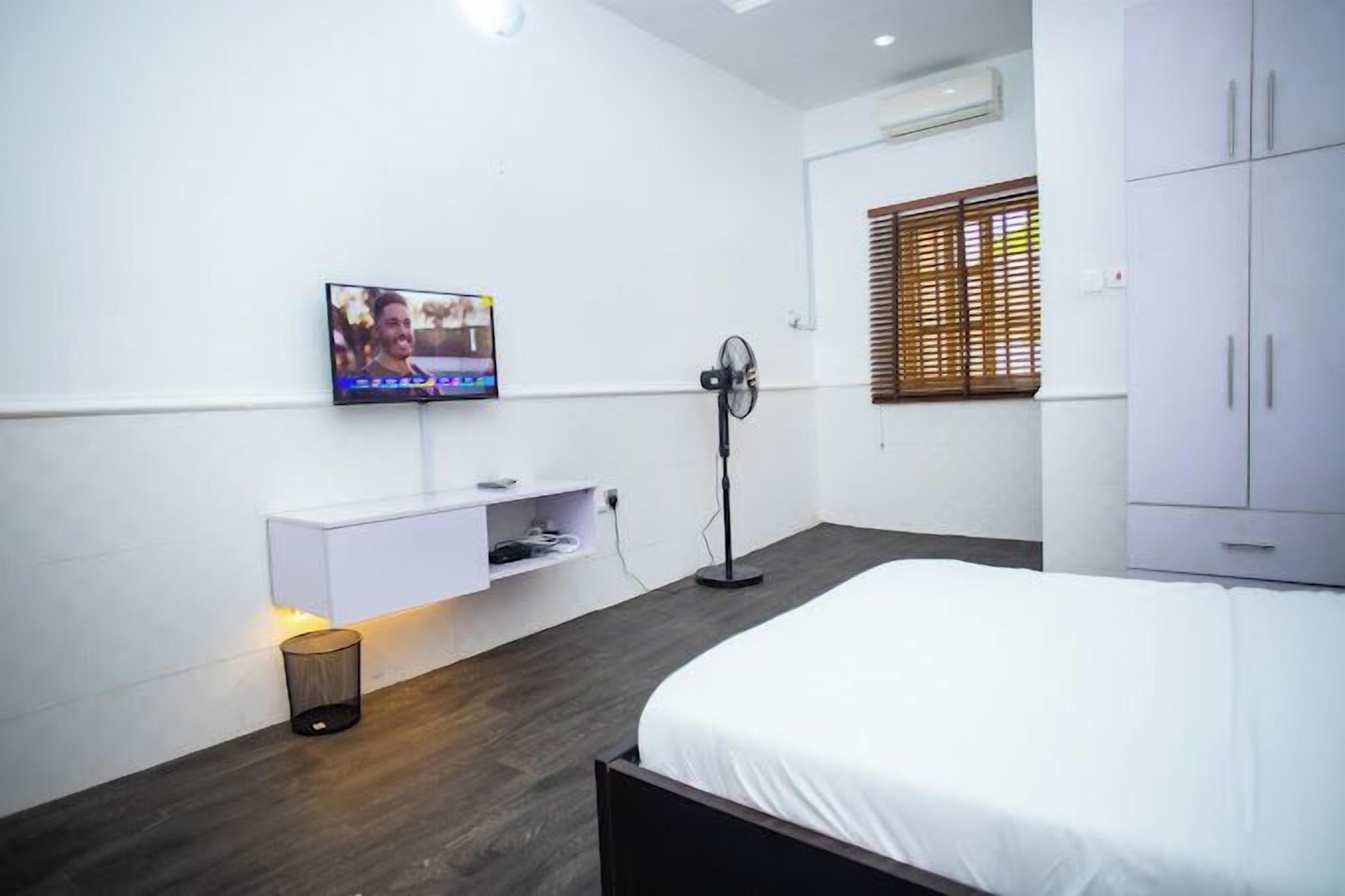 Una habitación de hotel moderna con una cama, un televisor montado en la pared, un ventilador de pie y un armario.