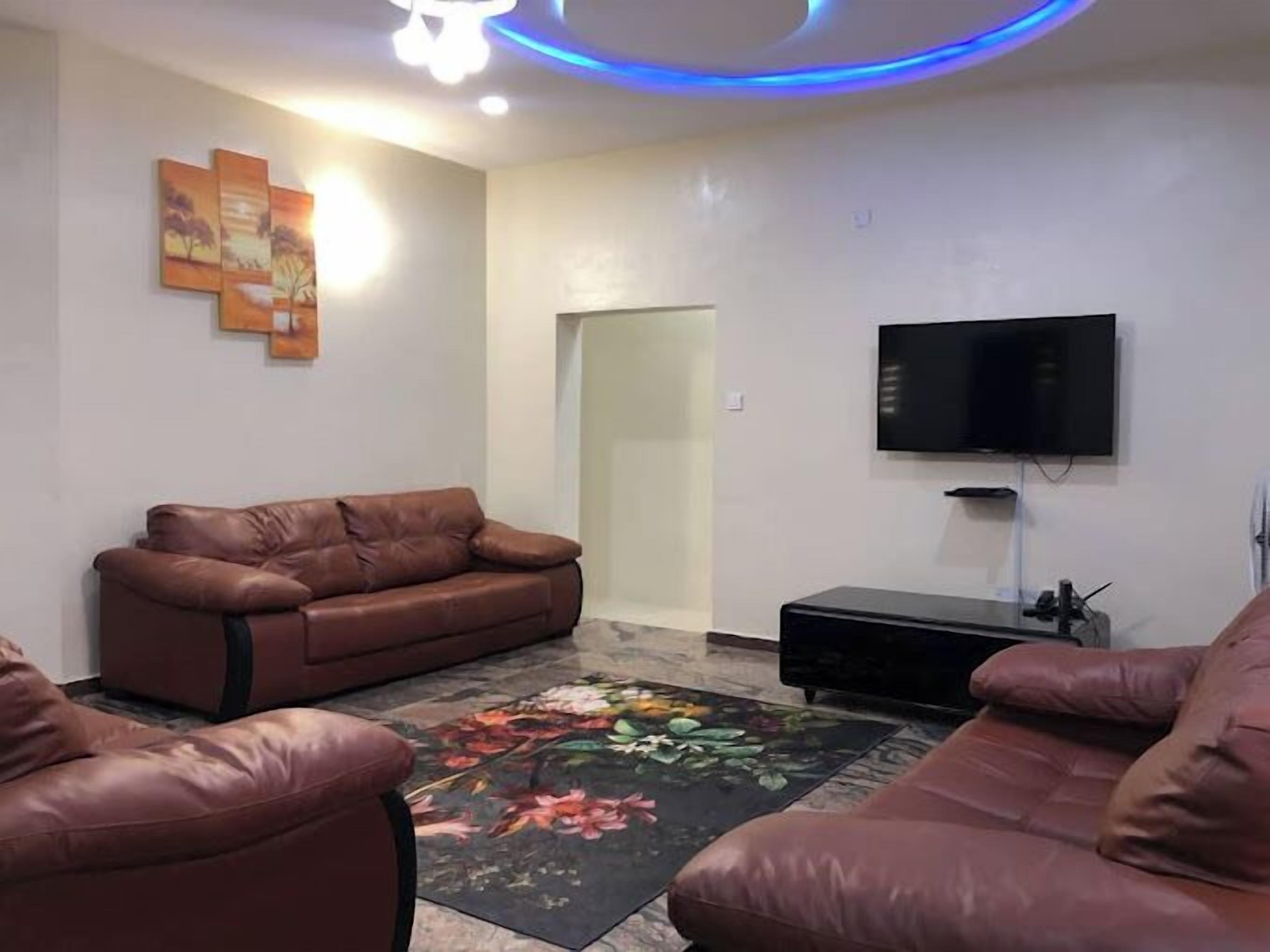 Un salon avec un canapé en cuir marron, un téléviseur à écran plat fixé au mur et un tapis à motifs floraux sur le sol.