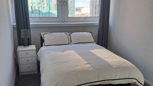A bedroom with a bed, a nightstand, a lamp, and a view of buildings through the window. - Luxury two bedroom skyline views apartment/Flat (Birmingham)