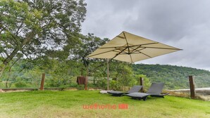 A grassy outdoor area with a large umbrella providing shade, two lounge chairs, and a view of a forested hill.