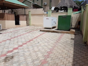 A paved courtyard with a thatched-roof structure, a white refrigerator, and a green utility box. - House Six Apartment (Abuja)