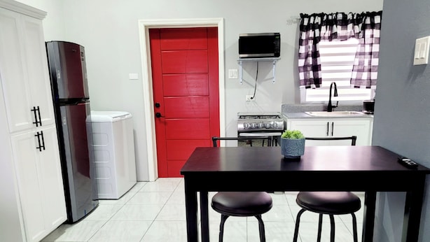 A compact kitchen with a red door, a microwave, a stove, and a dining table with two stools. - Rest Easy Suite #1 with Mountain and Sea Views (Spooners Village, Cayon)