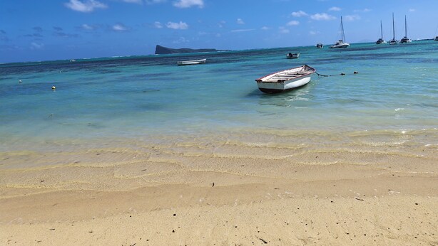 A beach with clear turquoise water, a sandy shore, and several boats anchored in the distance. - The nice corner (Grand baie)