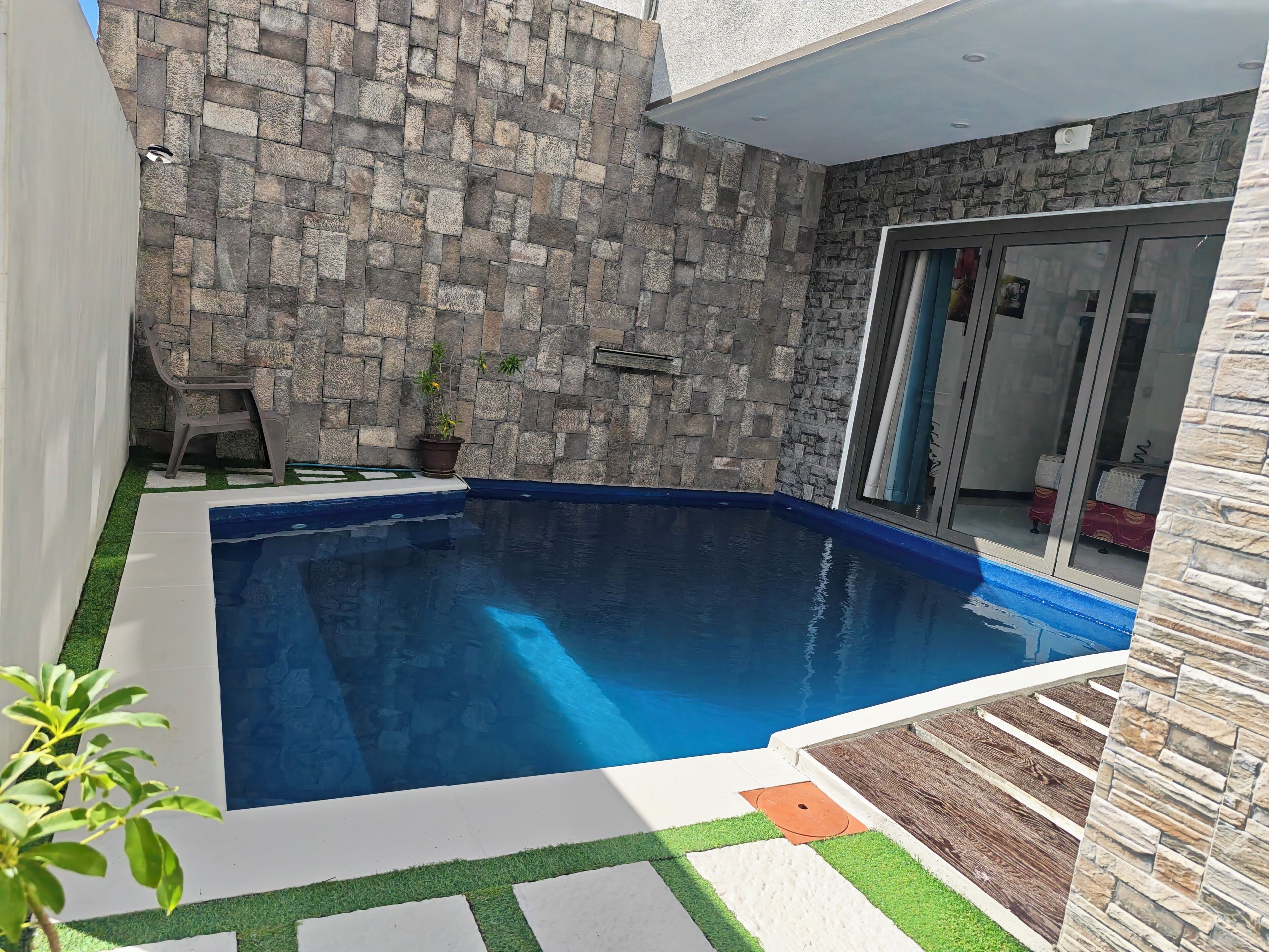 A swimming pool with a stone wall backdrop, a glass door entrance, and a small potted plant.