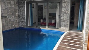 A swimming pool with a stone wall and glass doors leading to a room with a bed. - The nice corner (Grand baie)