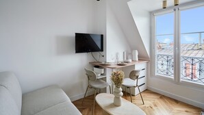 A modern living room with a TV, a dining table, chairs, and a large window with a view of buildings. - Superb Apartment Sacre Coeur With Air-conditionner (Paris)