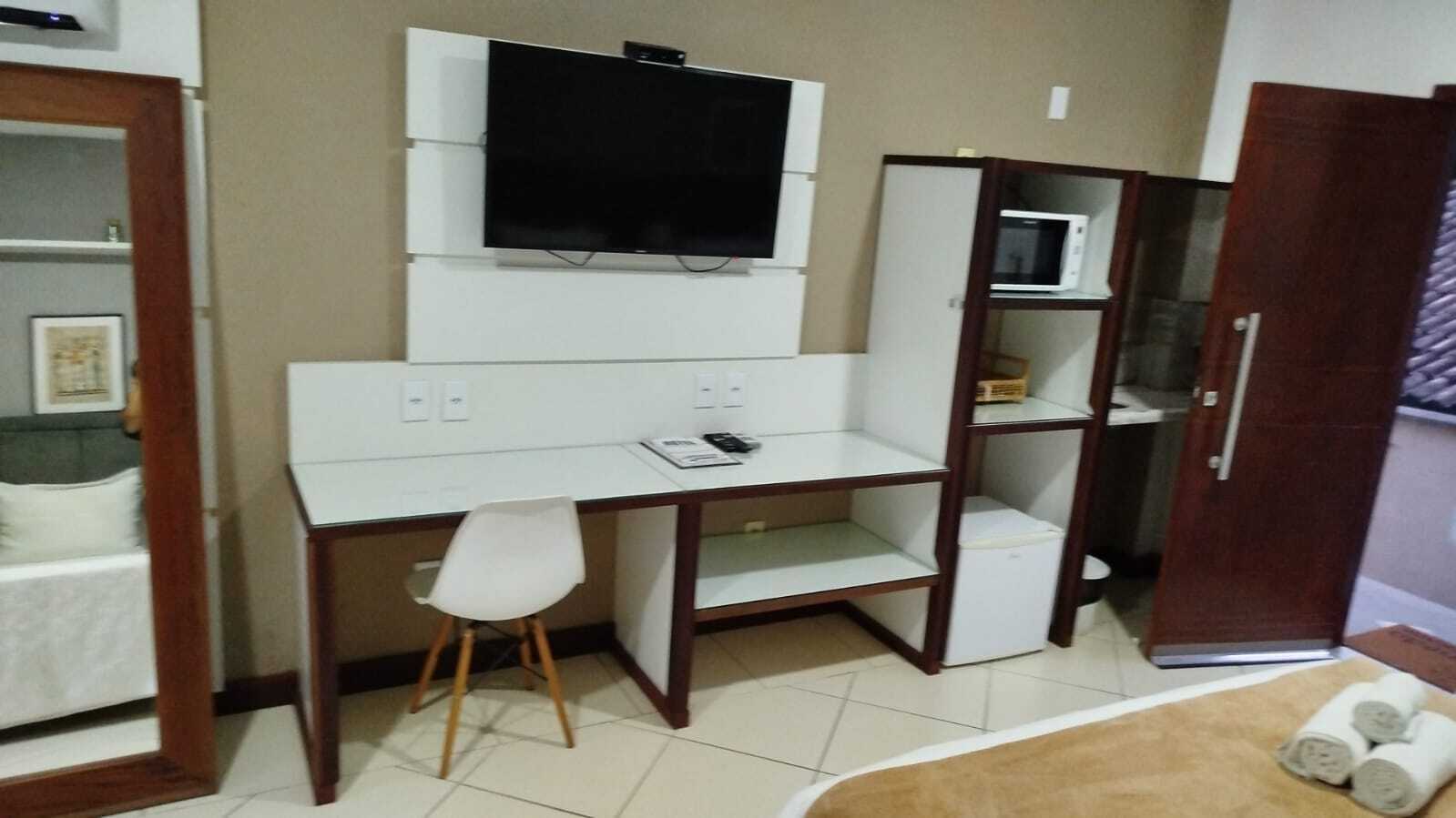 A compact room with a white desk, a chair, a television mounted on the wall, a microwave, and a wardrobe.