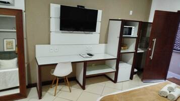 A compact room with a white desk, a chair, a television mounted on the wall, a microwave, and a wardrobe.