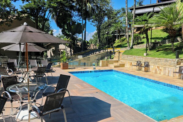 Piscina com guarda-corpo de vidro, rodeada por mobiliário de exterior com guarda-sóis e uma parede de pedra com vegetação.