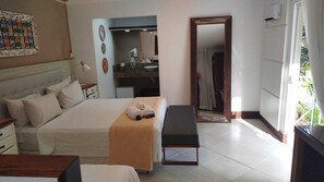 Quarto com cama, mesa de cabeceira, banco e banheiro visível por uma porta aberta.