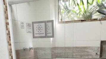 Zona de ducha con paredes de vidrio, piso de baldosas, un inodoro y una ventana con vista a plantas.