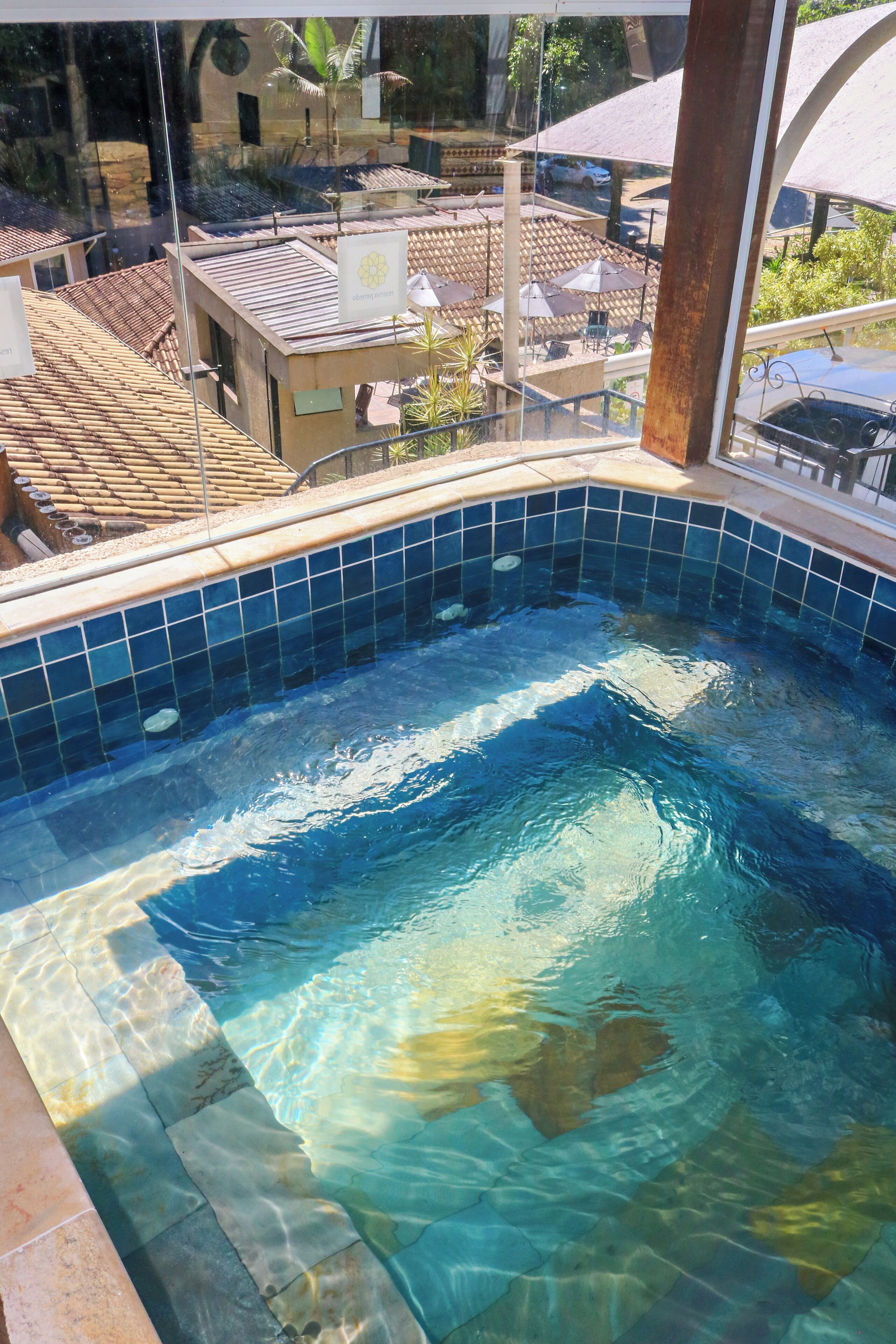 Una piscina con agua azul cristalina, bordes de azulejos y vistas del entorno, incluyendo edificios y sombrillas.
