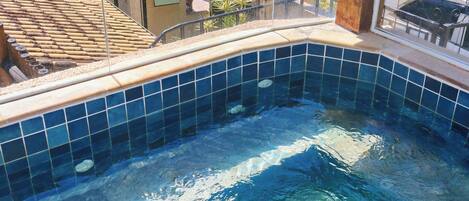 Una piscina con agua azul cristalina, bordes de azulejos y vistas del entorno, incluyendo edificios y sombrillas.