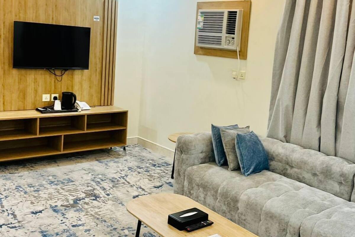A modern living room with a flat-screen TV, a wooden entertainment unit, a gray sofa with blue cushions, a coffee table, and a patterned rug.