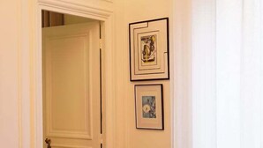A room with a brown leather armchair, a beige sofa with a patterned cushion, and a door leading to another room. There are two framed pictures on the wall.
