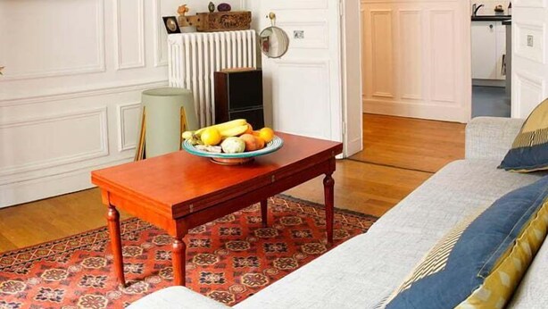 A living room with a sofa, a table with a fruit bowl, a radiator, and framed pictures on the wall.