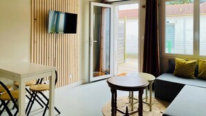 A modern living room with a TV mounted on a wooden wall, a sofa with yellow cushions, a small table, and a sliding glass door leading to a balcony. - Cocooning cottage by the sea  (Longeville-sur-Mer)
