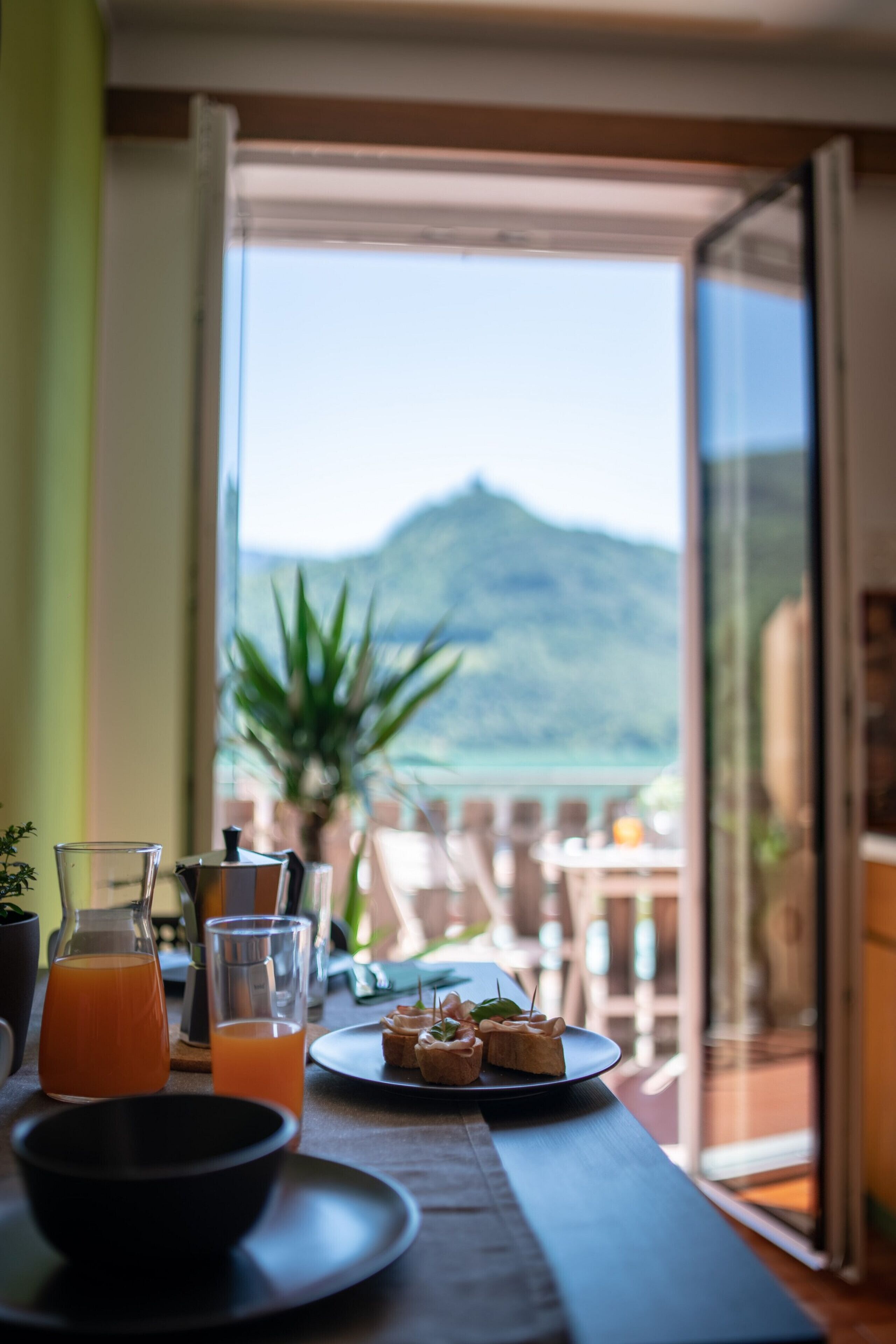 Une table à manger avec un verre de jus d’orange, une carafe et une assiette de hors-d’œuvre, le tout placé sur un balcon donnant sur une montagne.