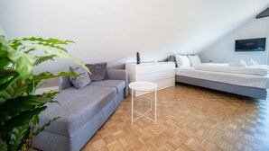 A modern bedroom with a grey sofa, a small white table, a bed with white bedding, and a TV mounted on the wall.
