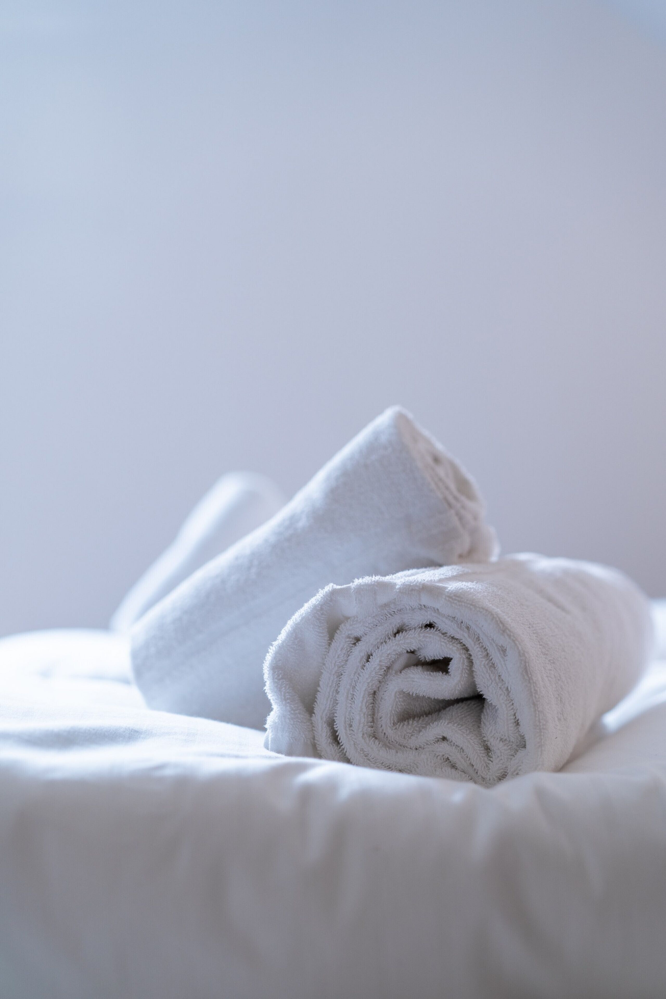 Une serviette blanche, soigneusement pliée, est posée sur un lit, le tout sur un fond uni.