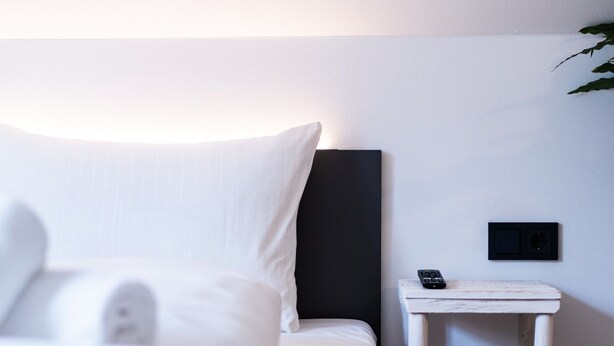 Una cama bien hecha con sábanas blancas y una almohada, una mesita de noche con un control remoto, y un enchufe en la pared.