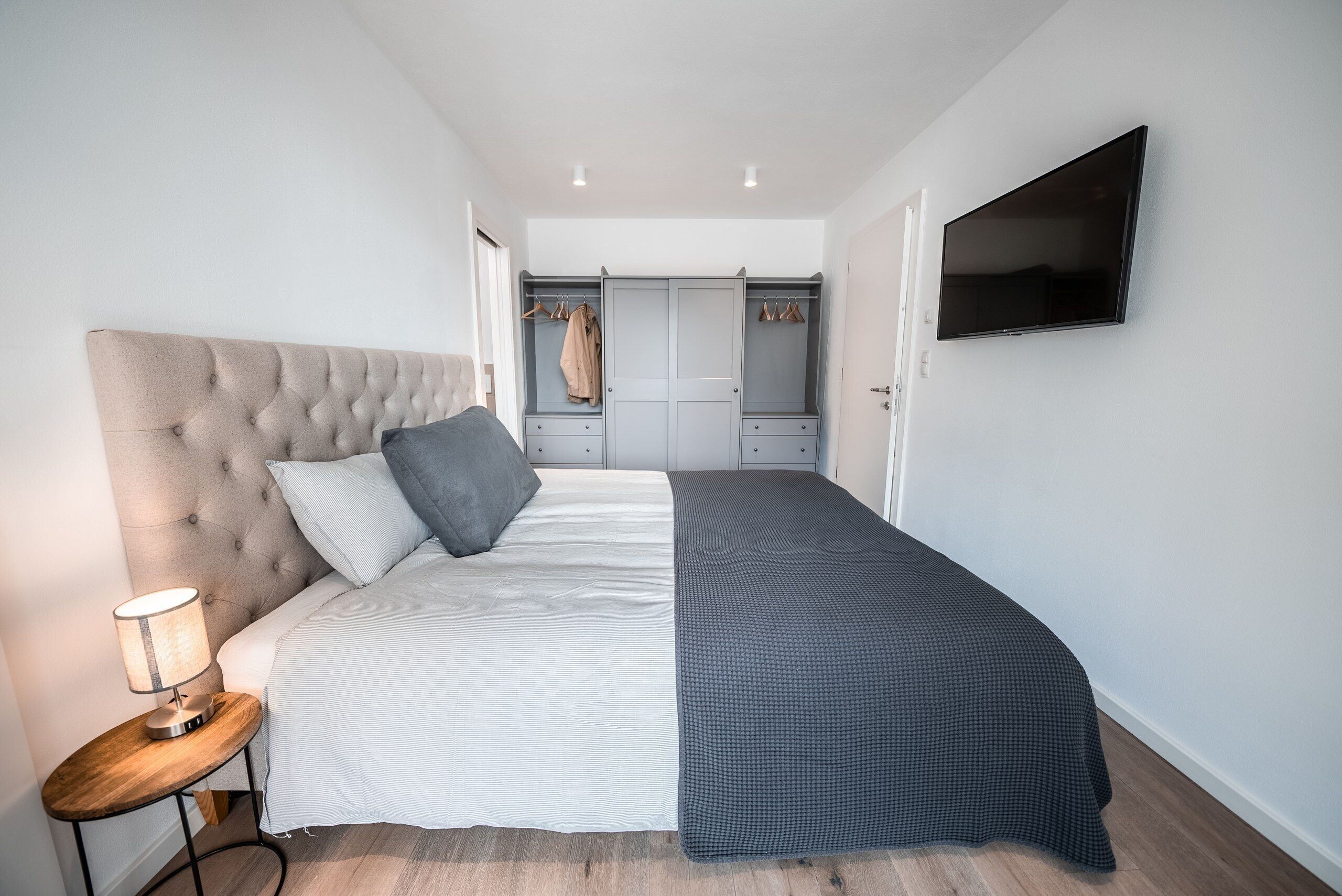 Un dormitorio con cama, mesita de noche, lámpara, armario y televisor montado en la pared.
