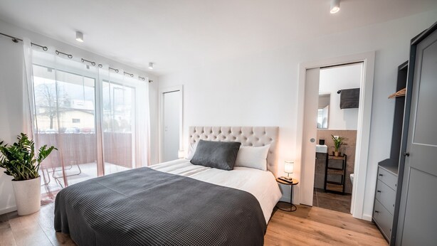 Une chambre avec un grand lit, une table de chevet avec une lampe, une porte-fenêtre coulissante donnant sur un balcon et une vue sur l’extérieur.