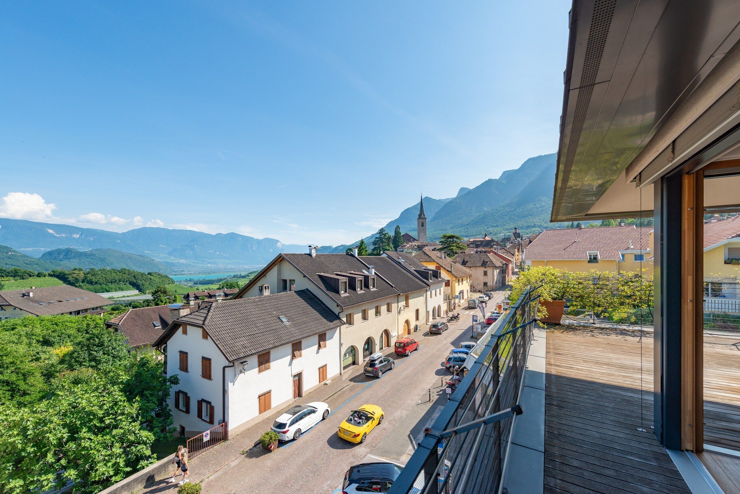 A balcony with a view of a mountain landscape, a street with parked cars, and traditional buildings.