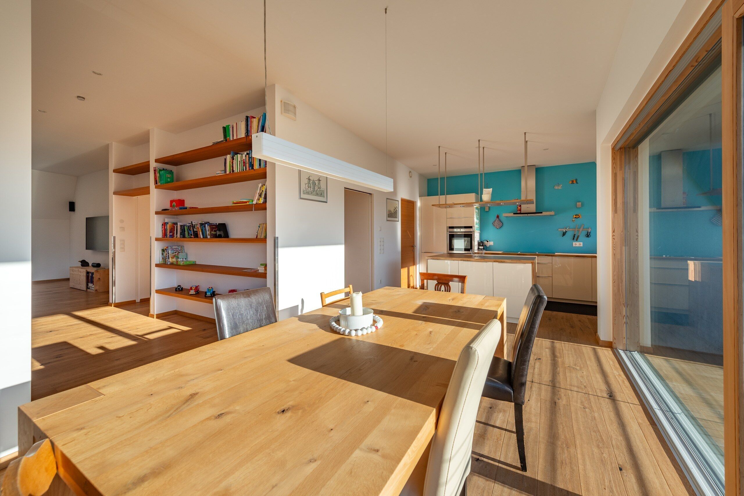 A modern kitchen with a blue accent wall, wooden shelving, and a dining area with a wooden table and chairs.
