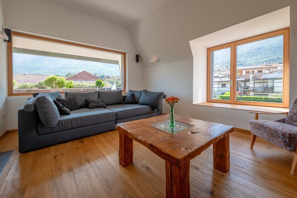 Una sala moderna con un sofá gris, una mesa de centro de madera y grandes ventanas que ofrecen vistas a un paisaje montañoso.
