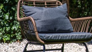 Una silla de mimbre con un cojín, sobre un suelo de grava y una mesa de cristal al frente.