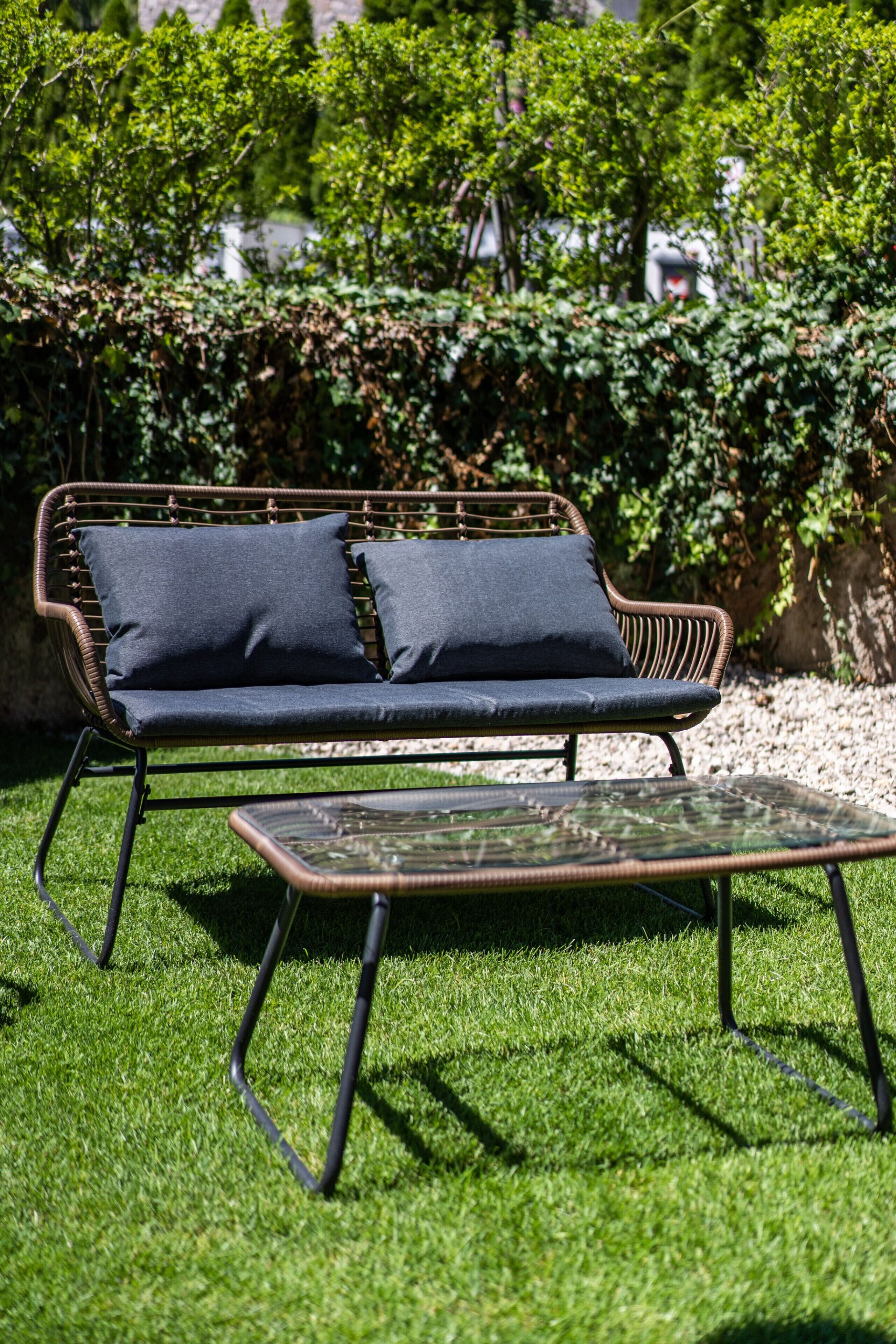 Un canapé en osier avec des coussins et une table basse en verre, le tout installé dans un espace extérieur verdoyant.