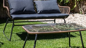 Un sofá de mimbre con cojines y una mesa de centro de vidrio en un área verde al aire libre.