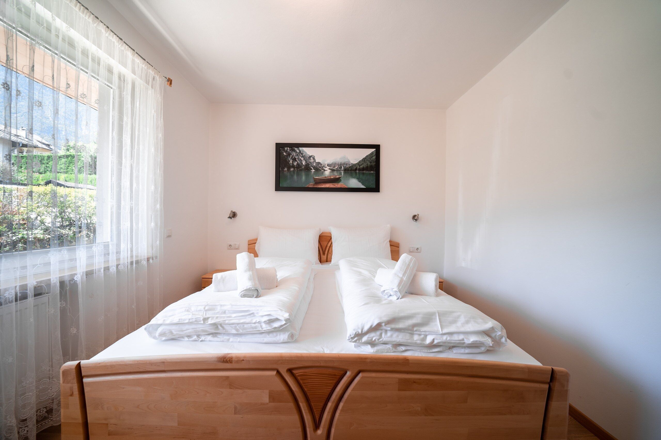 Una cama doble con ropa de cama y toallas blancas, cabecera de madera, ventana con cortinas traslúcidas y un cuadro en la pared.