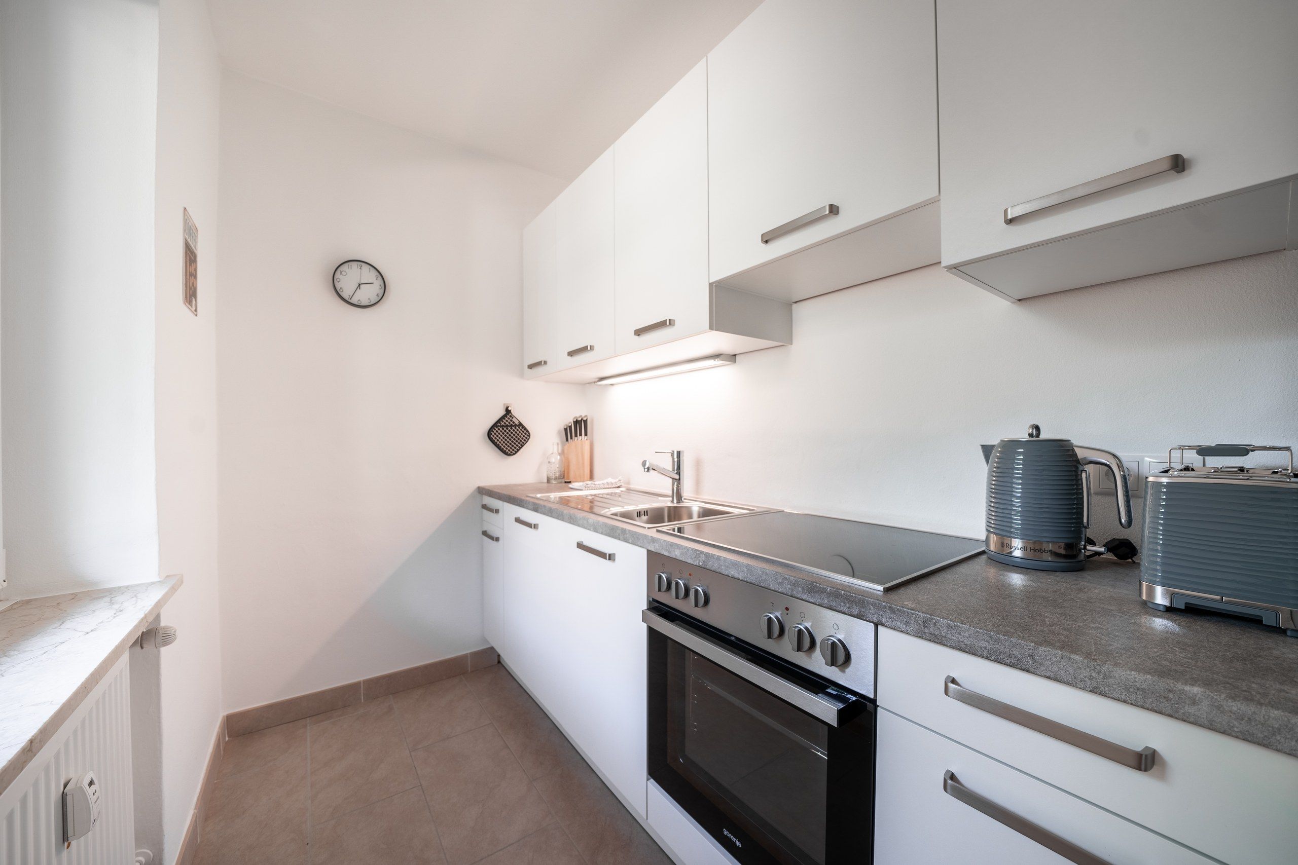 Cocina compacta con armarios blancos, horno integrado, cocina y hervidor sobre la encimera.