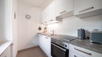 Cocina compacta con armarios blancos, horno integrado, cocina y hervidor sobre la encimera.