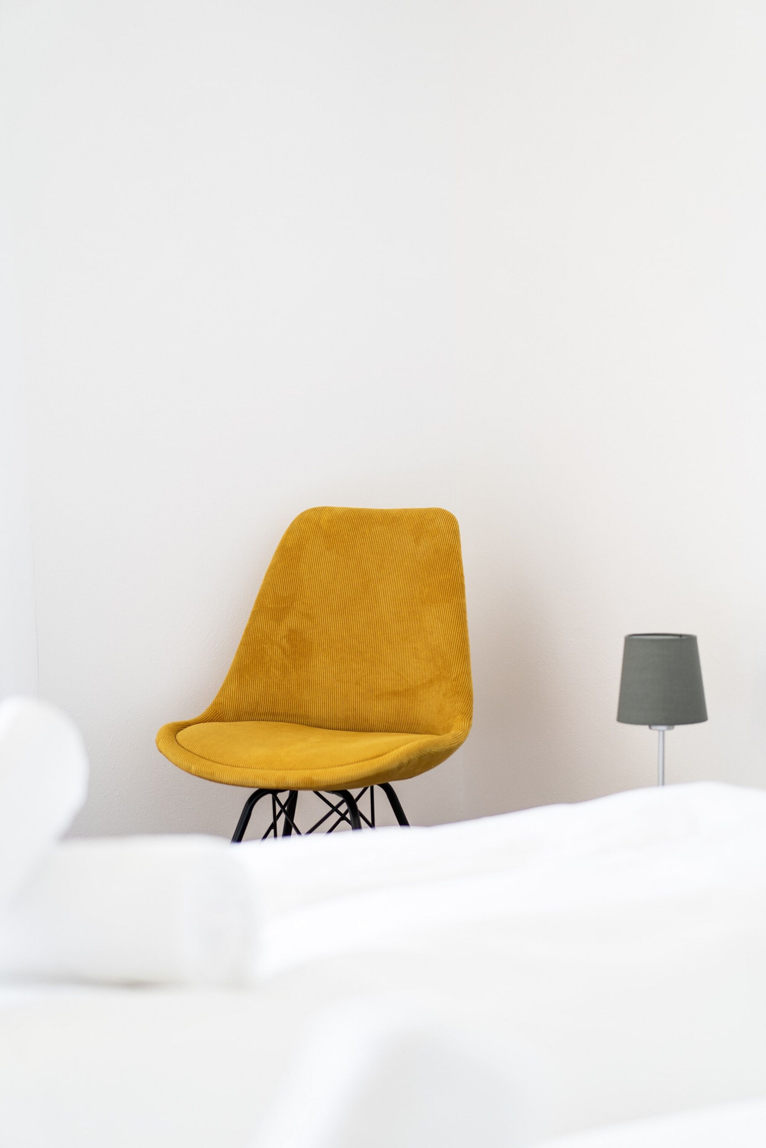 Una silla amarilla con textura acanalada, estructura de metal negro y una lámpara de pie gris frente a una pared blanca.