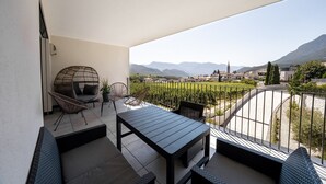Ein Balkon mit Tisch und Stühlen, einem Hängesessel und Blick auf einen Weinberg und ferne Berge.