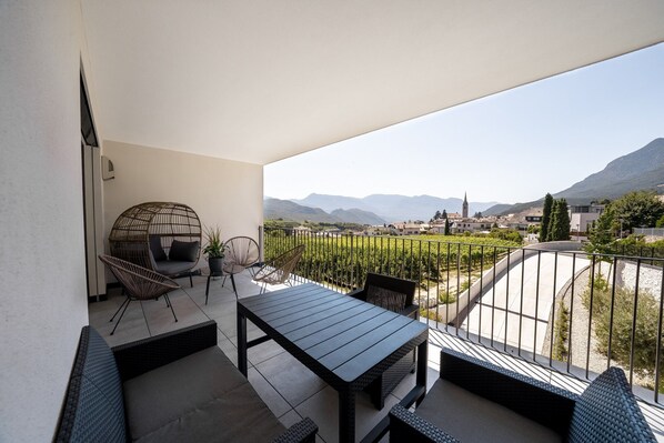 A balcony with a table and chairs, a hanging chair, and a view of a vineyard and distant mountains.
