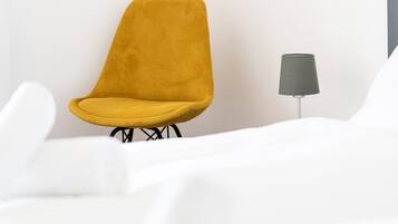Ein Zimmer mit einem einzelnen gelben Sessel, einem kleinen Tisch mit einer Lampe und weißer Bettwäsche.