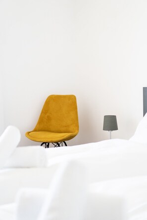 A room with a single yellow chair, a small table with a lamp, and white bedding.