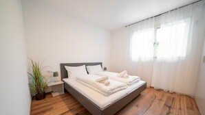 Ein modernes Schlafzimmer mit Holzboden, einem Bett mit weißen Bettwäsche, einem Nachttisch mit Lampe und einer Zimmerpflanze.